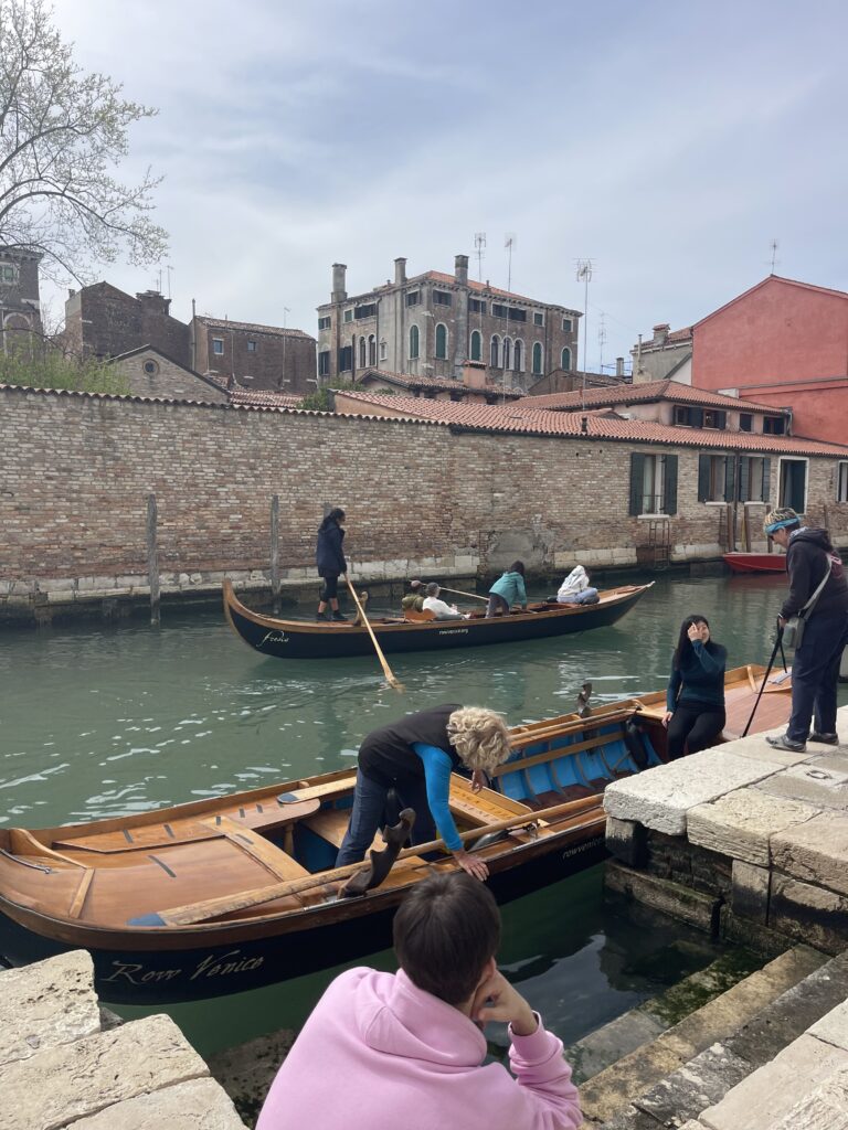 Group of people practicing voga alla veneta rowing technique with an instructor in a quiet Venice canal, as others watch from the stone edge while boarding traditional boats for a hands on rowing experience.