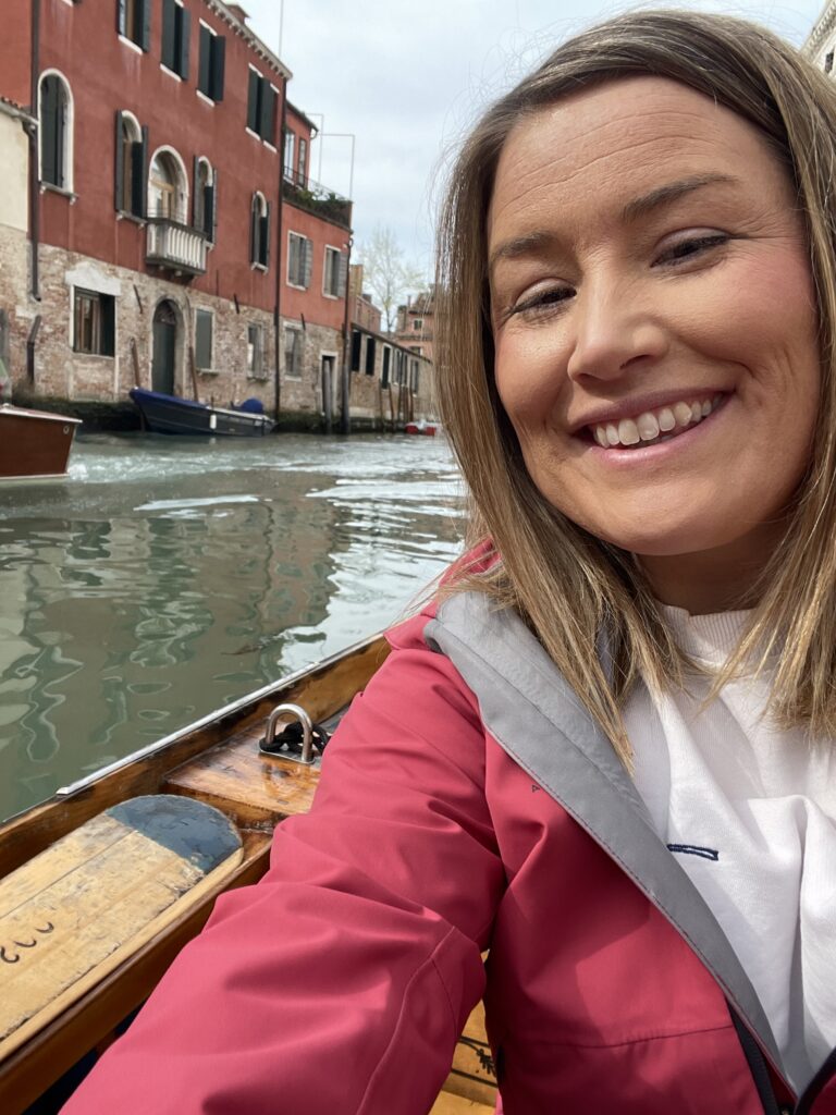 Smiling woman in a red jacket takes a selfie while standing in a wooden boat on a quiet Venice canal during a rowing lesson, with colorful historic buildings and moored boats lining the water behind her.
