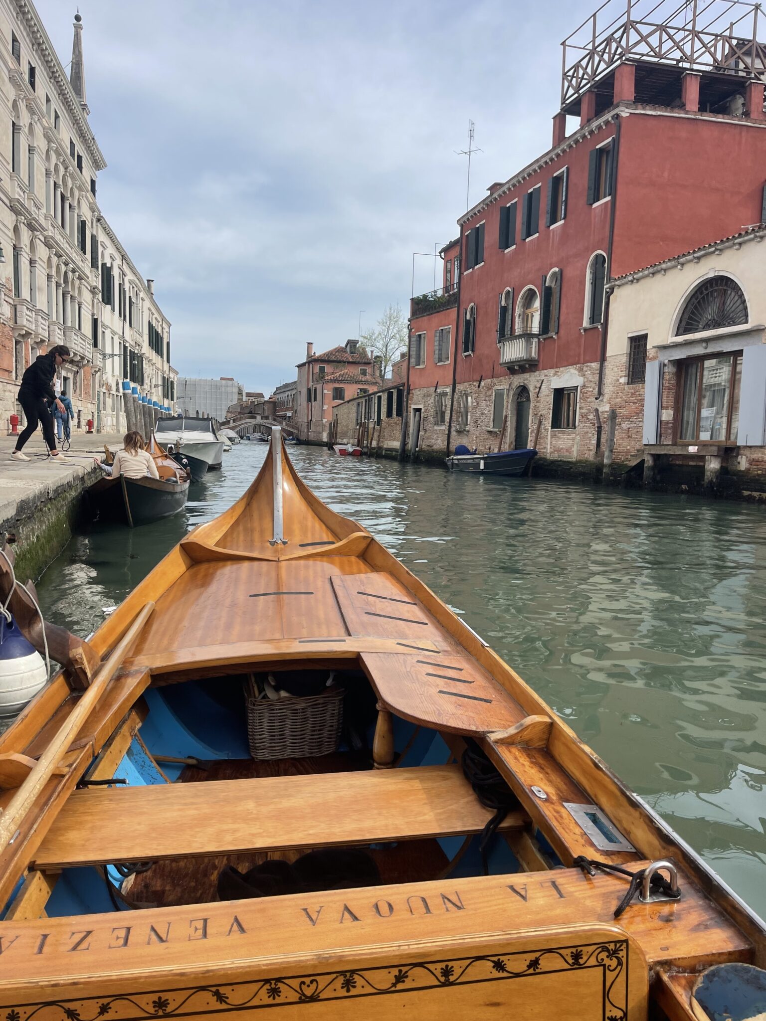 View from inside a wooden Venetian rowing boat looking down a narrow canal lined with red and cream buildings, showing the perspective during a rowing lesson in Venice as other boats pass by.