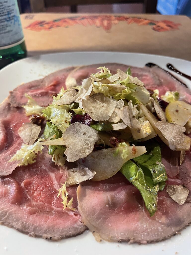 Thin slices of cured meat are topped with greens. pear. and shaved truffle on a white plate. This Tuscan meal shows the local food worth enjoying after a long day on the Via Francigena San Gimignano to Siena route.