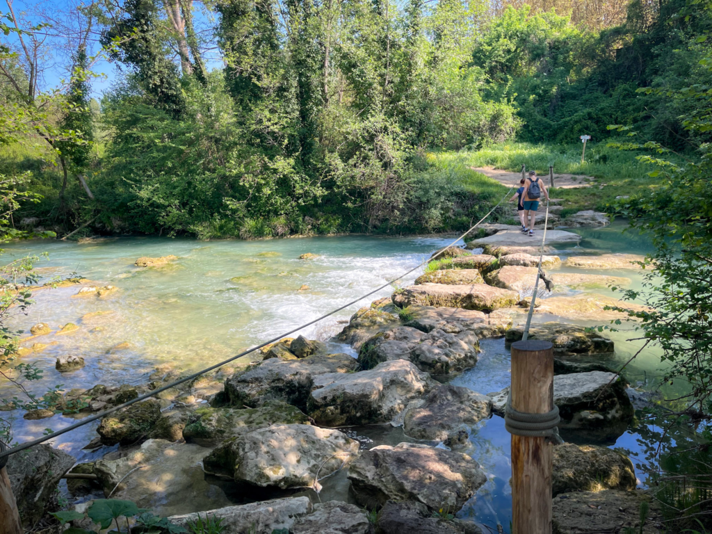 Two hikers cross a rocky river using stepping stones and a rope handrail through a lush green forest. The crossing shows one of the more adventurous sections of the Via Francigena San Gimignano to Siena walk at the Parco Fluviale Alta Val D'Elsa