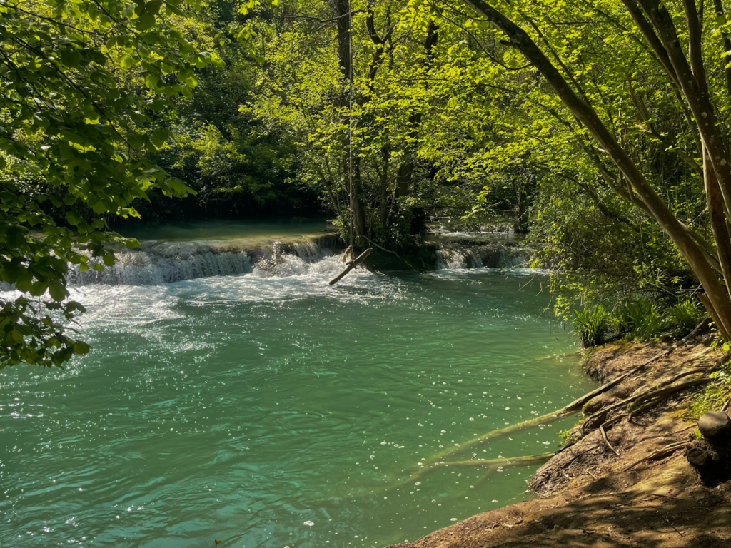 Turquoise water flows over small waterfalls in a shaded forest pool at Parco Fluviale Alta Val D’Elsa, with a rope swing hanging from the trees above. This peaceful river spot offers a scenic break near the Via Francigena San Gimignano to Siena route.