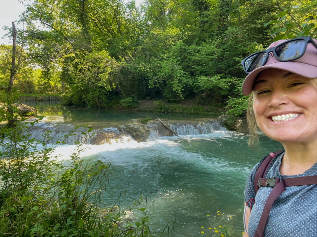 Smiling woman in a pink cap stands beside green vineyards and rolling Tuscan hills under dramatic clouds. The countryside view shows the landscape on the Via Francigena San Gimignano to Siena route.