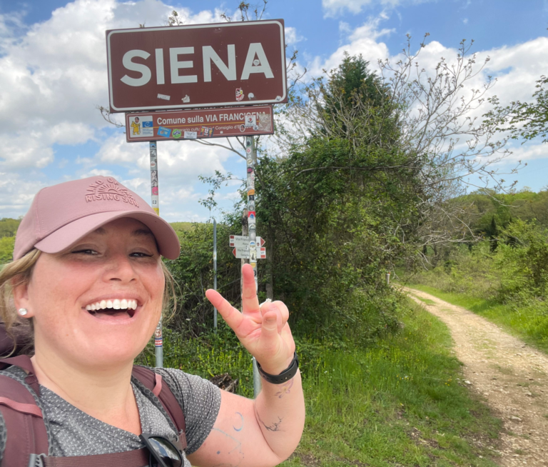 Smiling woman wearing a pink cap and backpack flashes a peace sign beside a large road sign reading “SIENA” with a smaller sign below that says “Comune sulla VIA FRANCIGENA.” A dirt path curves through green countryside under a partly cloudy sky, marking the final stretch of the Via Francigena San Gimignano to Siena route.