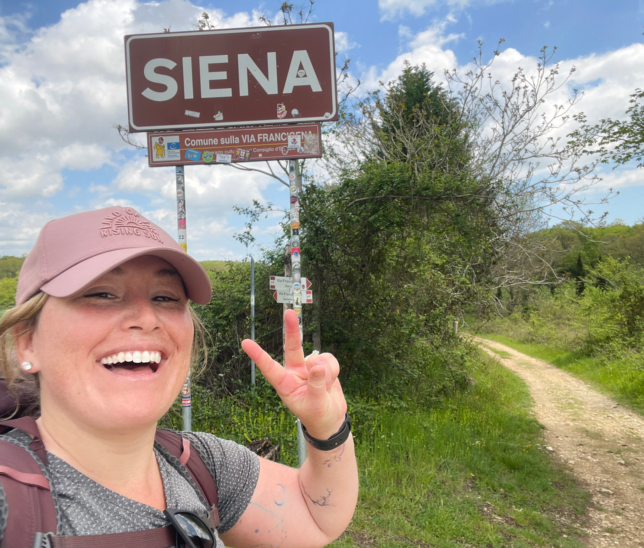 Smiling woman wearing a pink cap and backpack flashes a peace sign beside a large road sign reading “SIENA” with a smaller sign below that says “Comune sulla VIA FRANCIGENA.” A dirt path curves through green countryside under a partly cloudy sky, marking the final stretch of the Via Francigena San Gimignano to Siena route.