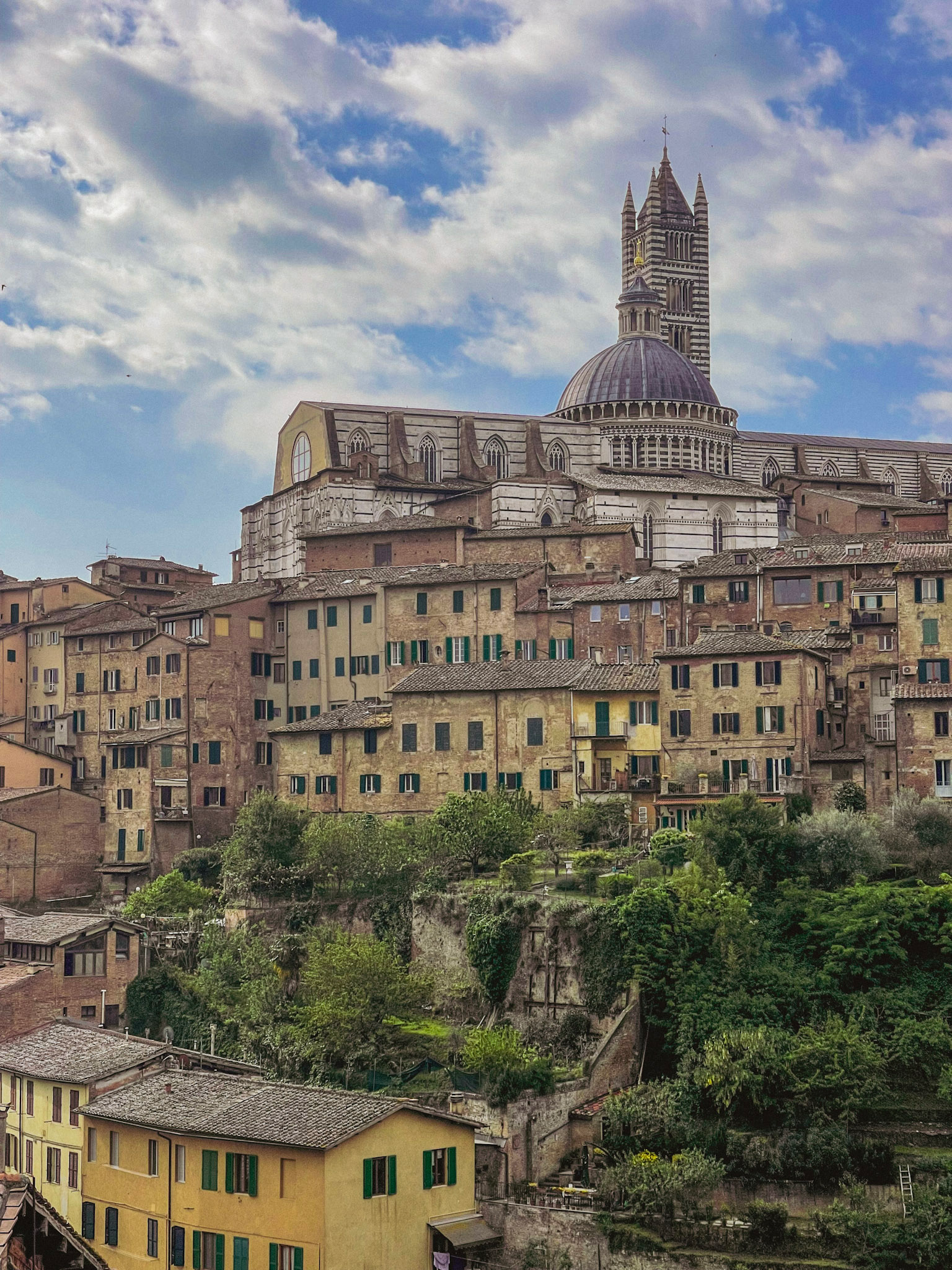 Siena’s cathedral dome and striped bell tower rise above tightly packed brick buildings and green shutters. The city skyline marks the destination after walking the Via Francigena San Gimignano to Siena route.