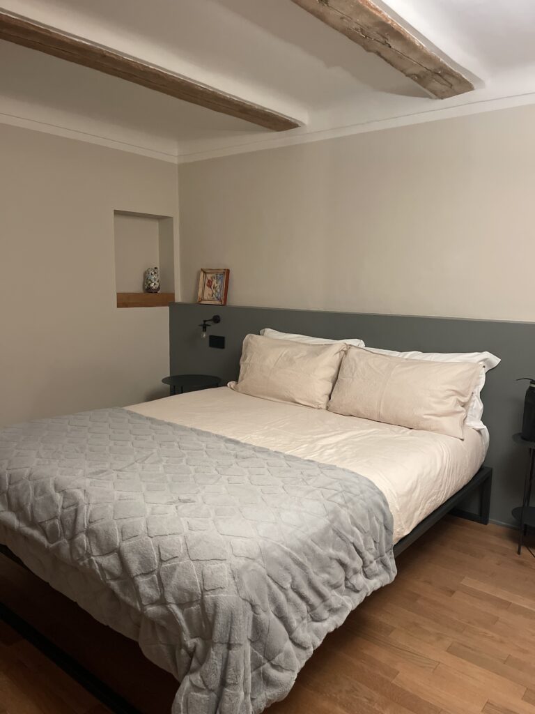 Minimalist bedroom at Casa Cicetta in San Marino with a large bed, neutral tones, wooden ceiling beams, and soft lighting offering a cozy stay during 2 days in San Marino.