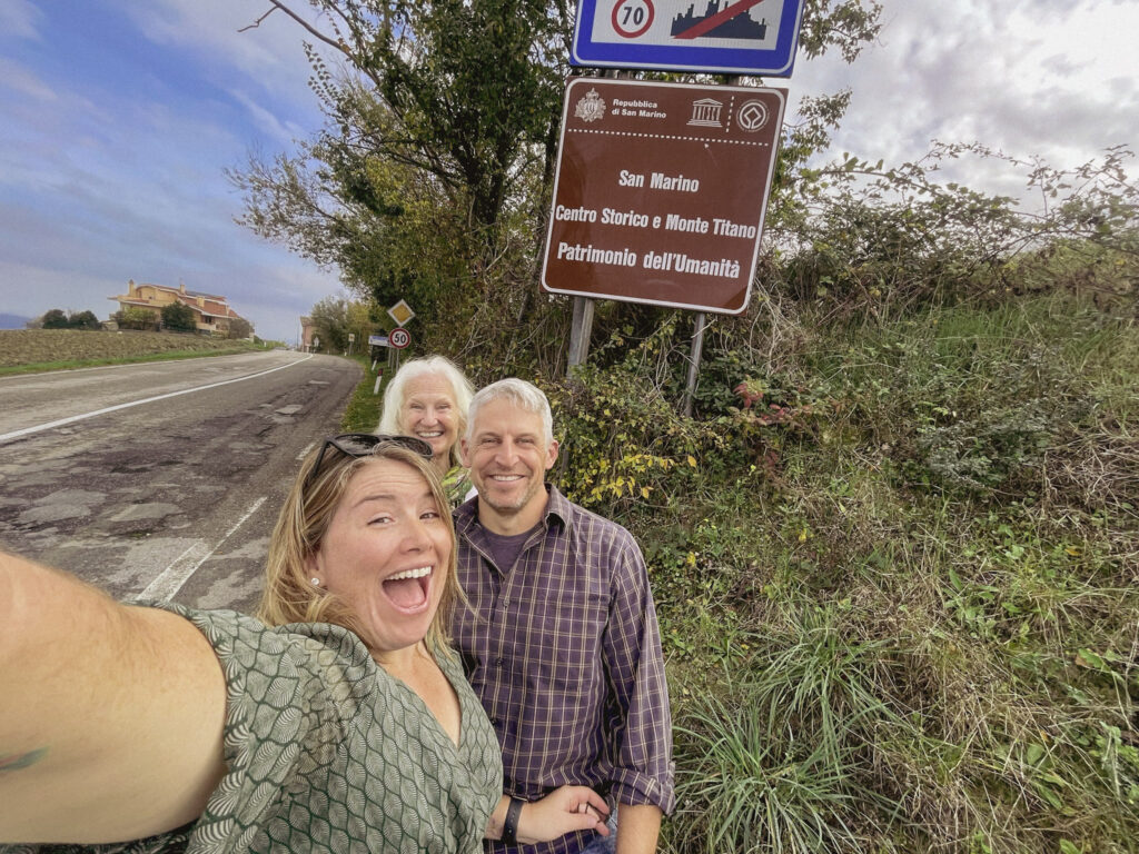 Three travelers smiling for a selfie beside a roadside sign that reads "San Marino Centro Storico e Monte Titano Patrimonio dell'Umanità" marking their arrival during a 2 days in San Marino itinerary.