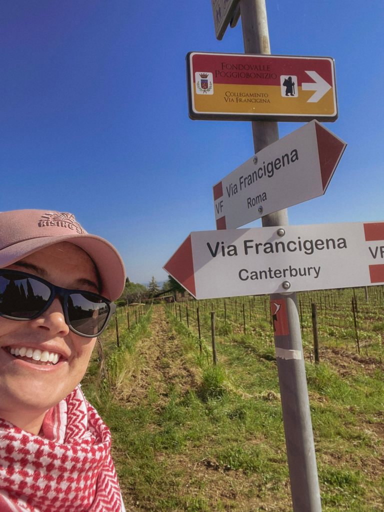 Smiling woman in a pink cap and sunglasses stands beside trail signs reading “Via Francigena Roma” and “Via Francigena Canterbury” in a vineyard. The signs point in opposite directions and mark the route through the Tuscan countryside between San Gimignano and Siena.