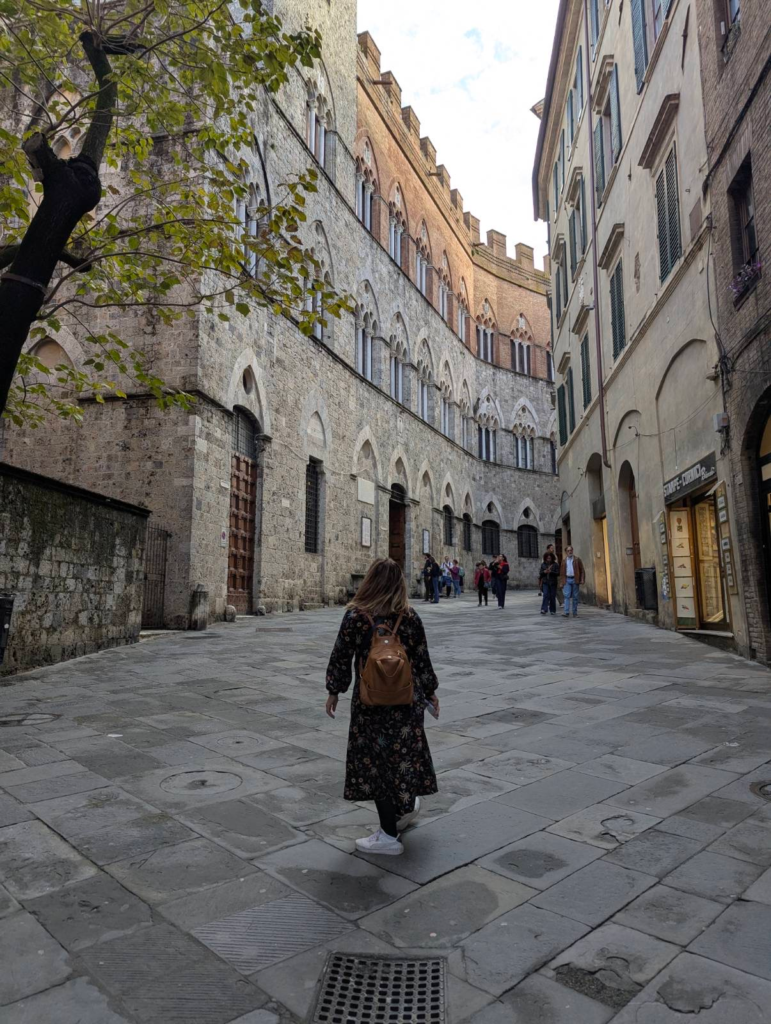 Woman with a backpack walking through a narrow stone street in Siena lined with historic buildings and arched windows. The curved facade and lively atmosphere capture everyday moments you might experience on a 2 days in Siena itinerary.