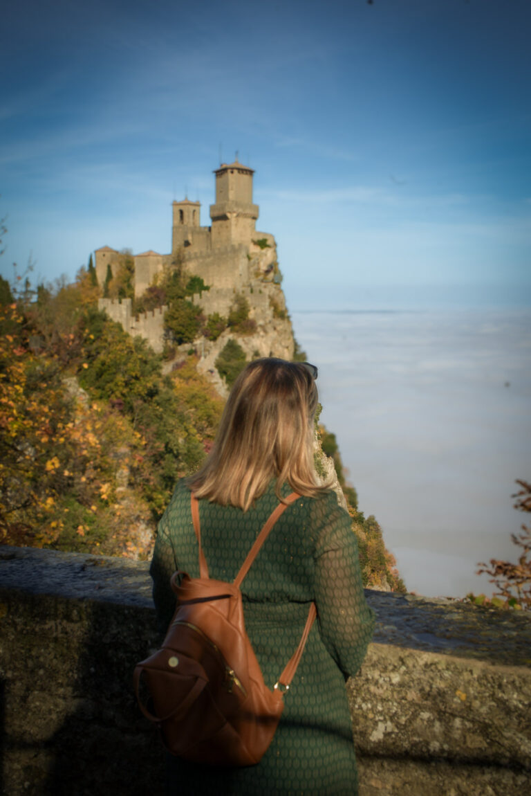 A woman with a brown backpack looks out toward a medieval fortress on a cliff above the clouds, a scenic viewpoint to experience during 2 days in San Marino.