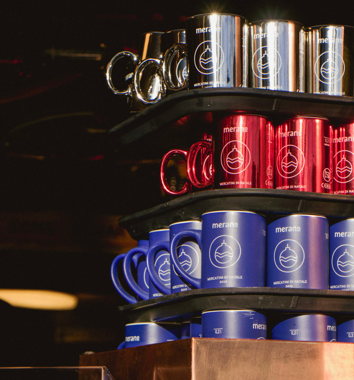 Stacked souvenir mugs in silver, red, and blue on display at the Merano Christmas Market, each labeled with “Merano” and “Mercatino di Natale.” Below the mugs, a copper stand features an apple graphic and bilingual text reading “Mela calda” and “Heisser Apfel,” indicating hot apple drinks.