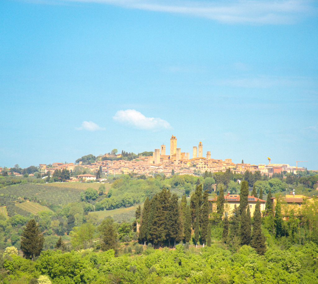 San Gimignano rises on a Tuscan hilltop with its medieval stone towers visible above green vineyards, cypress trees, and countryside. This scenic view shows a landmark stop along the Via Francigena San Gimignano to Siena route.