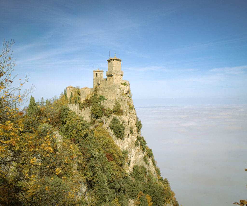 Guaita Tower perched on a rocky cliff atop Monte Titano surrounded by golden autumn trees and overlooking a thick layer of clouds, a signature view to experience during 2 days in San Marino.