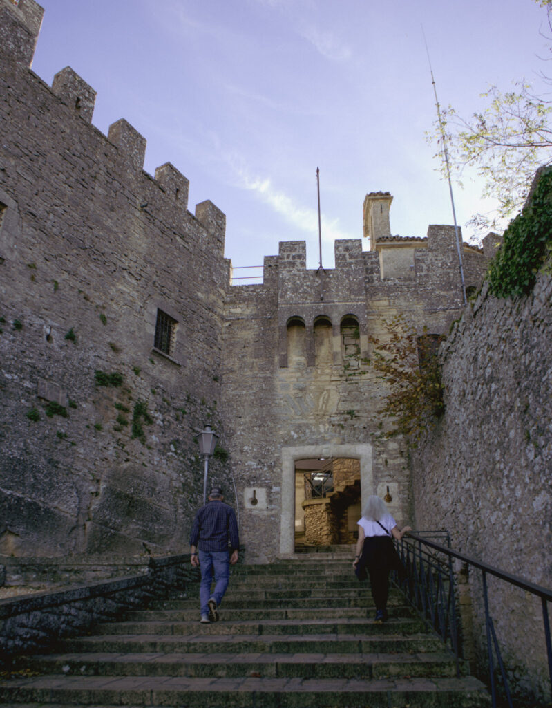 Two visitors walk up stone steps toward a medieval fortified gate with high stone walls and battlements, entering the historic center during 2 days in San Marino.