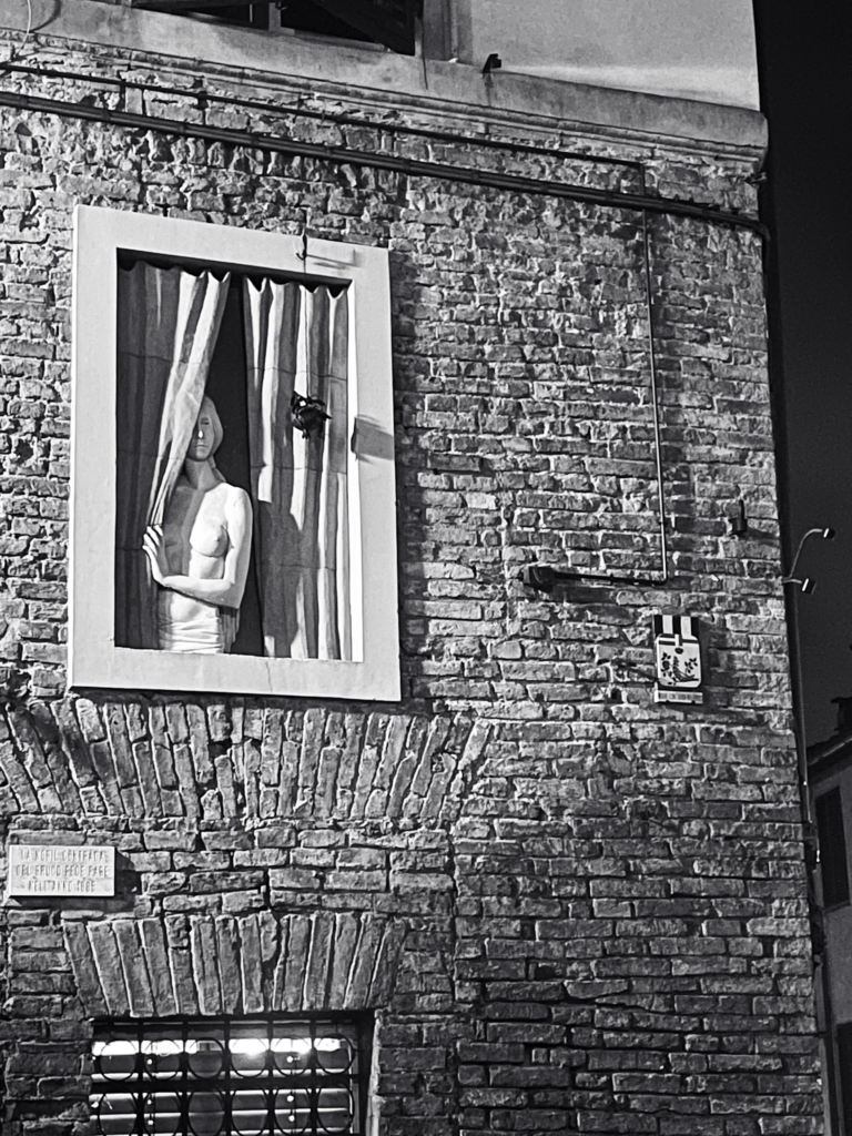 Black and white photo of a brick building in Siena with a painted mural of a woman pulling back curtains in a window frame. The street art contrasts with the historic stone walls, adding a modern artistic touch to a 2 days in Siena itinerary.