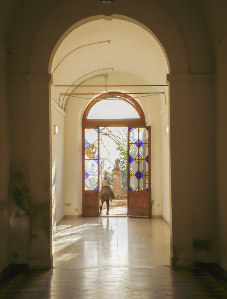 Softly lit arched hallway in Siena leading to open wooden doors with stained glass panels as a person steps outside into the evening light. This quiet transition scene captures the start of a dusk walk during a day 2 Siena itinerary.