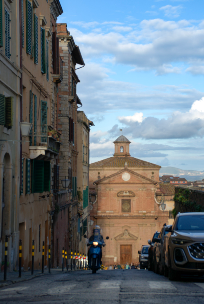 loping street in Siena with pastel buildings and green shutters leading down to a small church with a dome. A scooter drives toward the viewer adding movement to this everyday city scene on a 2 days in Siena itinerary.