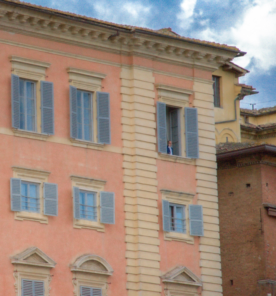 A man stands at an open blue-shuttered window of a pink palazzo in Siena, surrounded by closed shutters and stone trim.