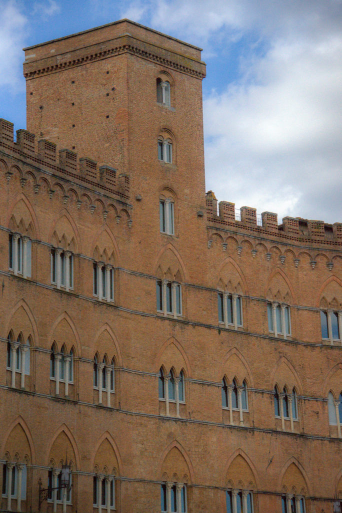 A medieval Sienese tower rises against blue sky above rows of pointed Gothic arched windows in warm brick.