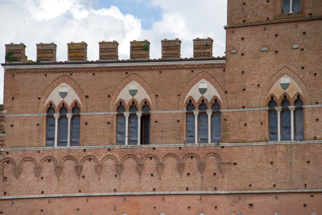 Close detail of Gothic arched windows and faded contrade shields on the brick facade of a Sienese palazzo.