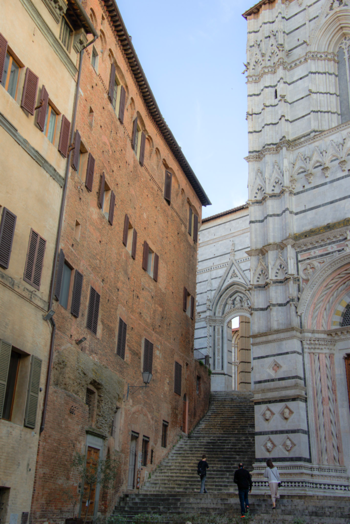 A stone staircase climbs between brick palazzos in Siena toward the striped marble facade of the Duomo cathedral.