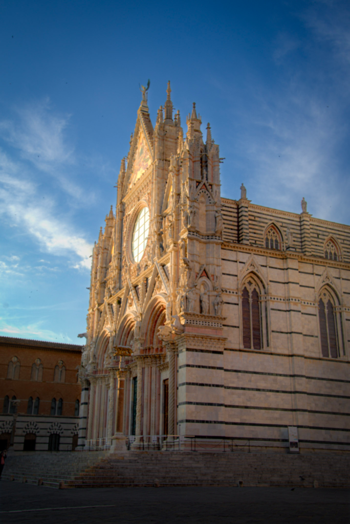 The full Gothic facade of the Siena Duomo with its rose window, sculpted saints, and ornate spires against blue sky.