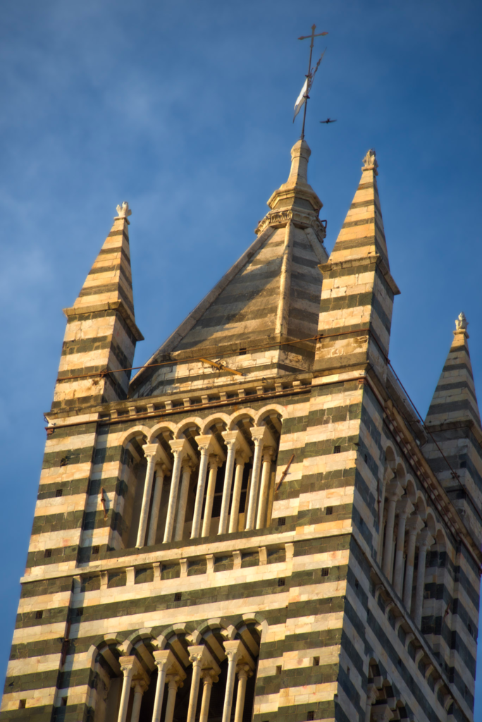 The striped marble bell tower of the Siena Duomo seen from directly below, with sharp spires against a darkening blue sky.