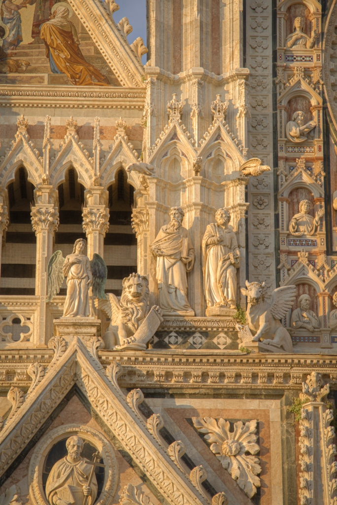 Cluster of sculpted saints, a winged lion, and a winged ox on the upper facade of the Siena Duomo, gilded by late afternoon sun.