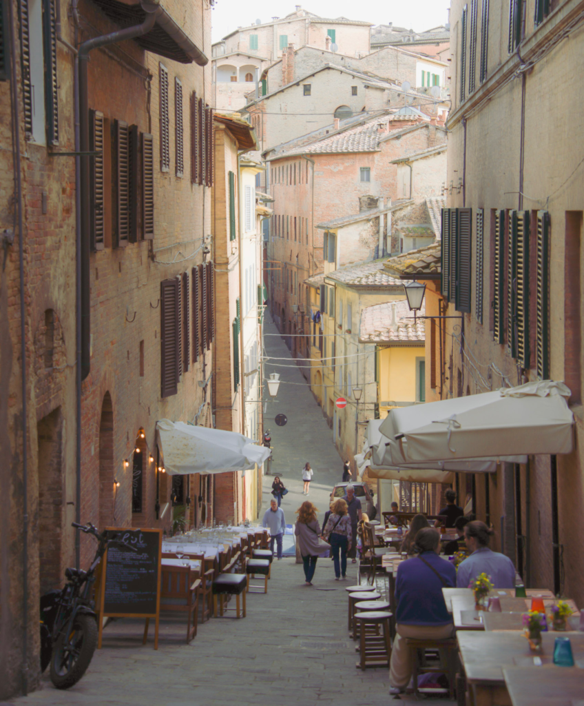 Outdoor restaurant tables line a sloping cobblestone alley in Siena leading down toward the historic city center.
