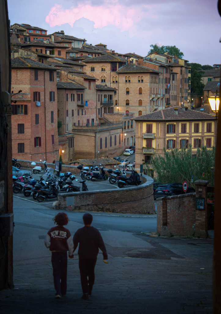 Two people walk downhill toward a cluster of historic Siena buildings at dusk with scooters parked along a curved stone wall and soft pink clouds in the sky. This scenic moment captures the relaxed pace of an evening stroll in Siena on day 2.
