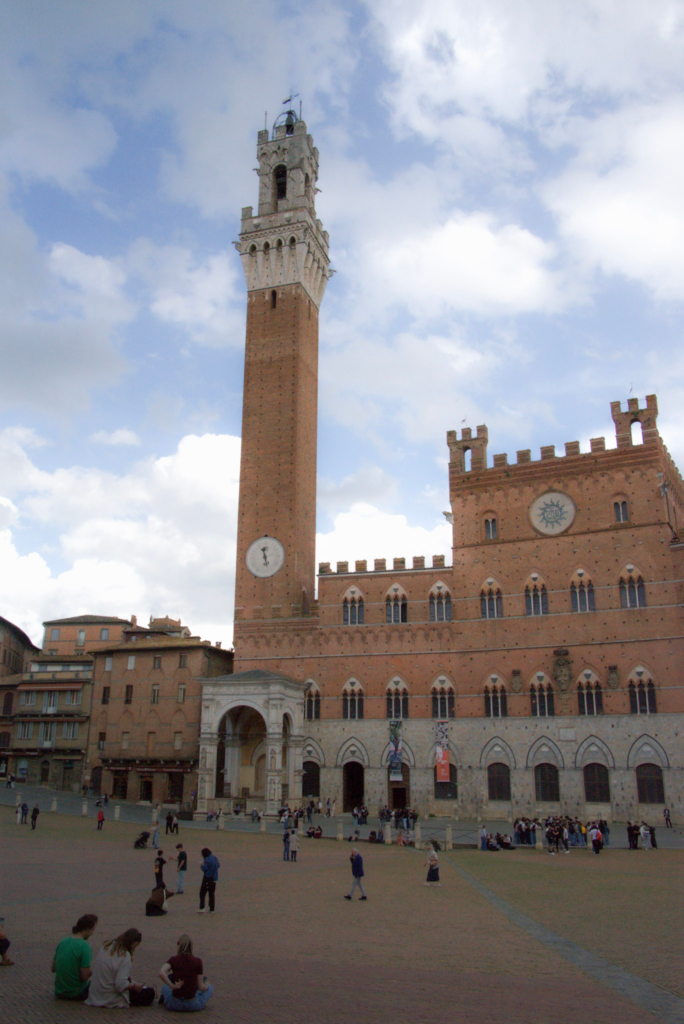 The Torre del Mangia and Palazzo Pubblico rise above Piazza del Campo by day with people scattered across the warm brick paving.