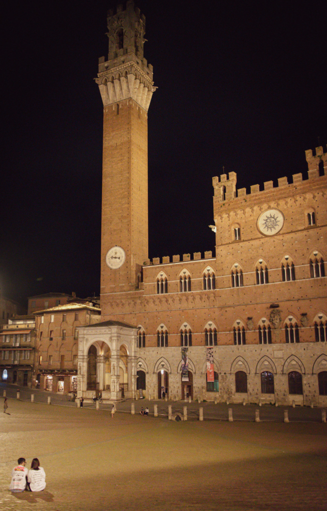 Piazza del Campo at night with Torre del Mangia and Palazzo Pubblico illuminated against a dark sky while a few people sit quietly in the square. This peaceful evening view highlights a must see stop on a Siena dusk walking itinerary.