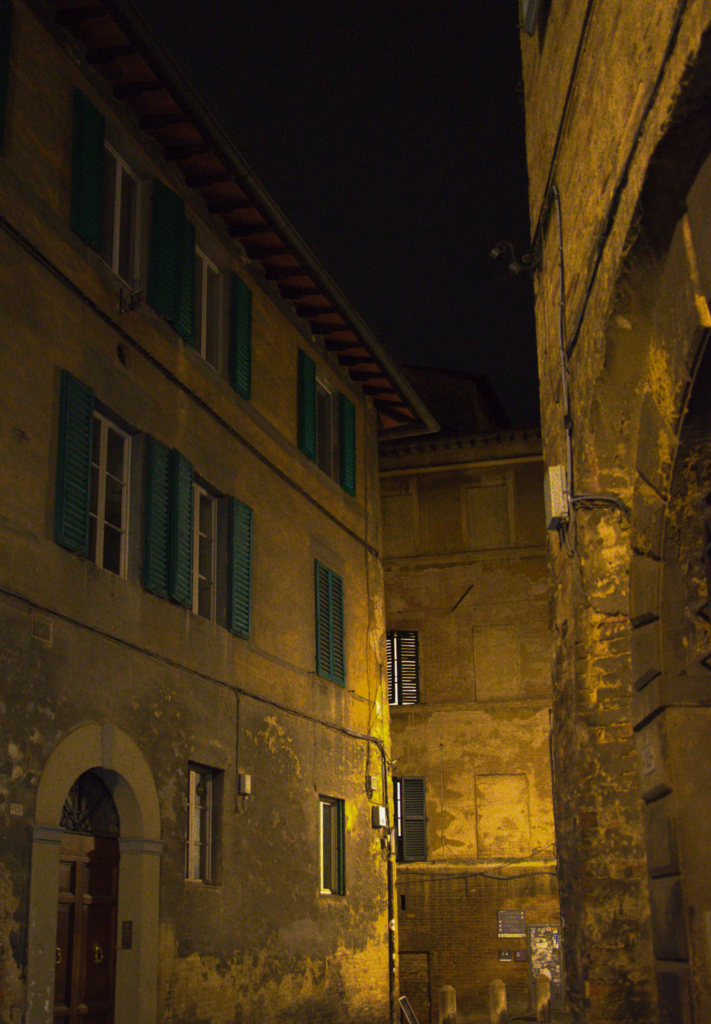 Narrow Siena street at night with warm yellow lighting casting shadows on textured stone walls and shuttered windows. This atmospheric alley reflects the charm of exploring Siena after sunset on day 2.