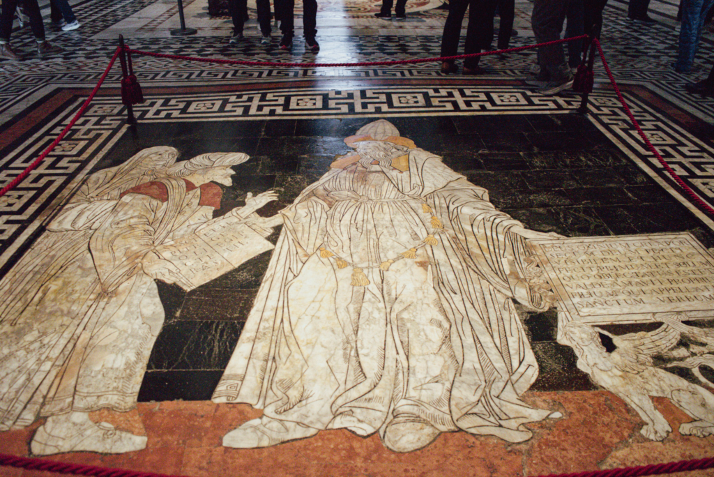Marble floor mosaic showing robed figures holding detailed panels with engraved text. The intricate floor designs are a highlight of the Siena Duomo and feature storytelling through art.