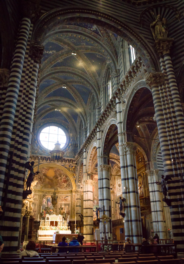 Expansive view of the Siena Duomo interior with rows of arches and striped columns leading toward the altar. The ceiling is decorated with stars creating a sense of depth and grandeur.