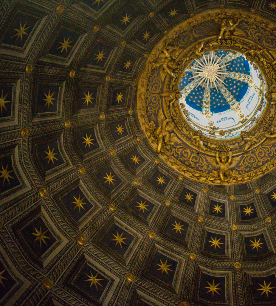 Dome ceiling filled with gold stars arranged in geometric patterns around a central circular opening. The rich decoration draws the eye upward inside the Siena Duomo.