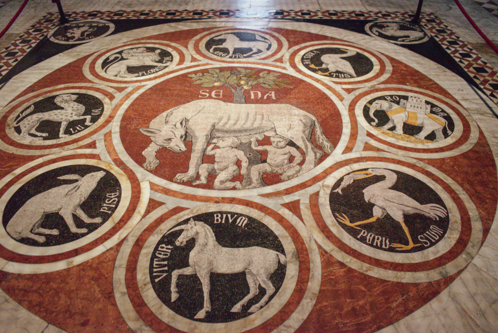 Circular marble mosaic showing animals arranged around a central scene of a wolf nursing two infants. The symbolic design is one of the many intricate floor pieces in the Siena Duomo.