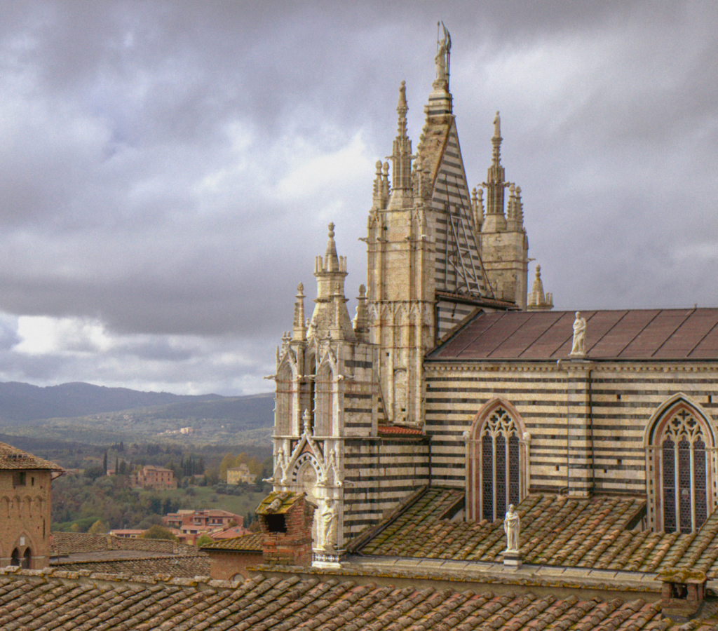 Side view of the Siena Duomo with its black and white striped marble facade and ornate spires rising above terracotta rooftops. Rolling Tuscan hills stretch into the distance under a cloudy sky, a view often seen on a 2 days in Siena itinerary.