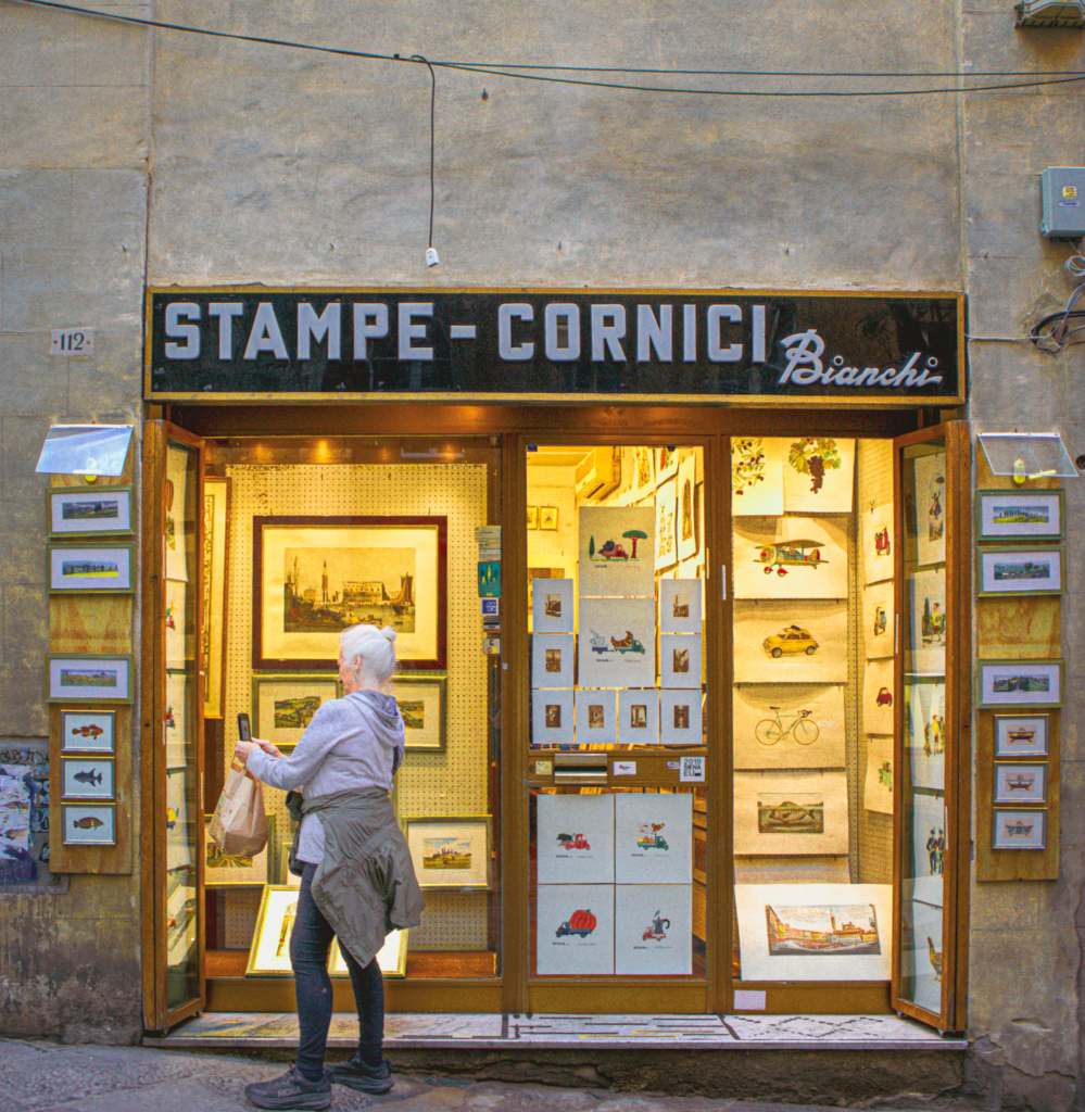 Woman standing outside a small art shop in Siena with the sign "STAMPE - CORNICI Bianchi" above the entrance. Framed prints and illustrations fill the doorway, capturing a charming shopping moment on a 2 days in Siena itinerary.