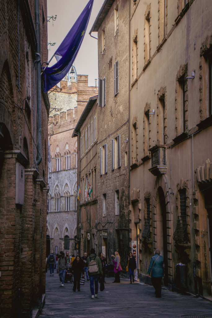 Narrow Siena street lined with tall historic stone buildings and shuttered windows as people walk along the cobblestone path toward a Gothic facade in the distance. A blue flag hangs overhead adding color to this lively daytime scene on a Siena itinerary.
