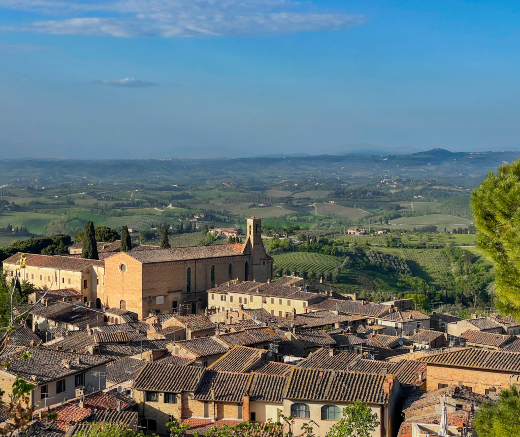 Terracotta rooftops and a brick church overlook rolling Tuscan hills, vineyards, and cypress trees from a high viewpoint in San Gimignano. The sweeping countryside view shows the landscape surrounding the Via Francigena San Gimignano to Siena route.
