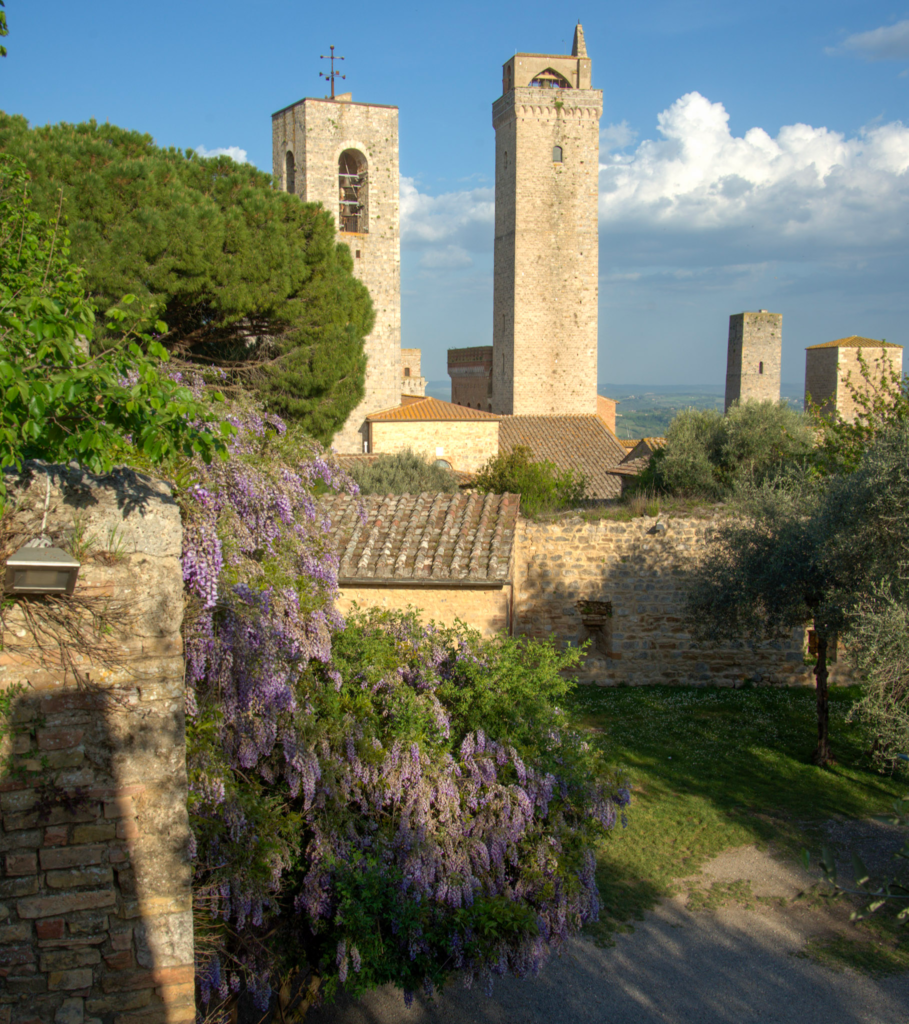 Purple wisteria spills over old stone walls below San Gimignano’s towers and rooftops. A quiet garden path curves through the historic hilltop town.