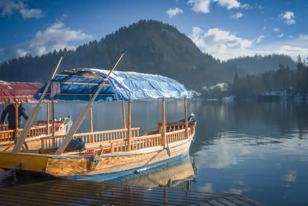 Traditional wooden pletna boat at Lake Bled dock in winter with snow on canopy and Julian Alps in background