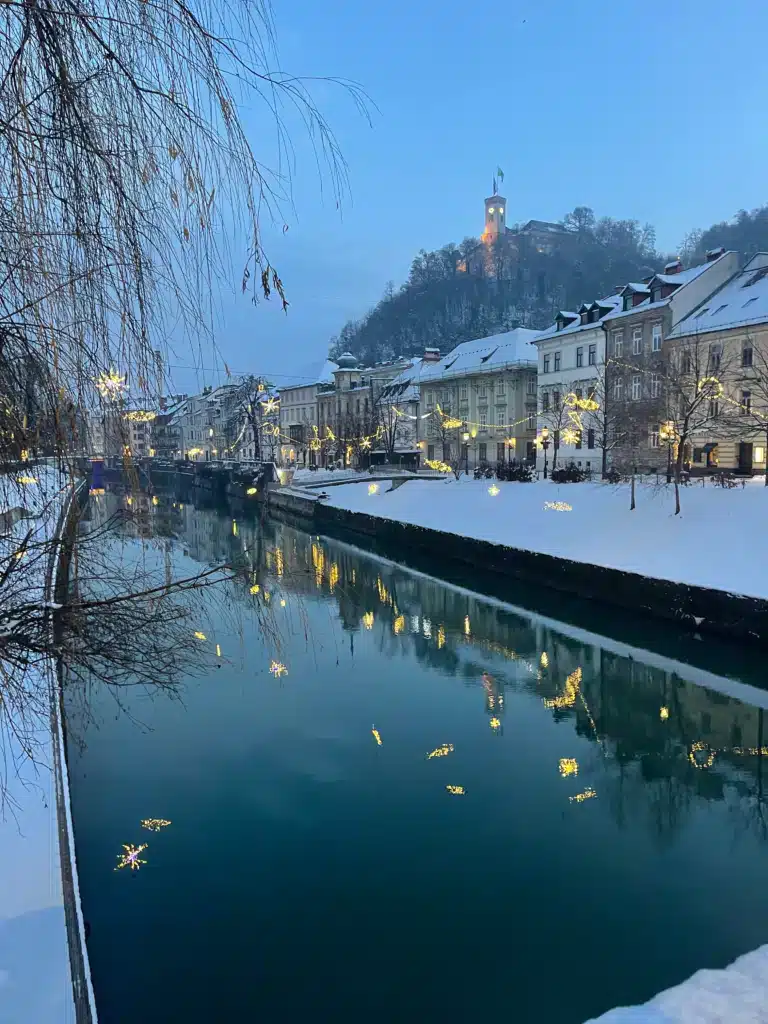 Ljubljana old town in winter at dawn with snow, Christmas lights reflected in the Ljubljanica river, and Ljubljana Castle on the hill