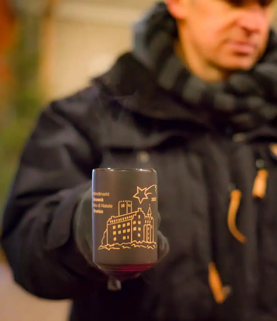 The 2025 Bruneck Christmas Market souvenir mug with a gold castle and shooting-star design, held up with mulled wine