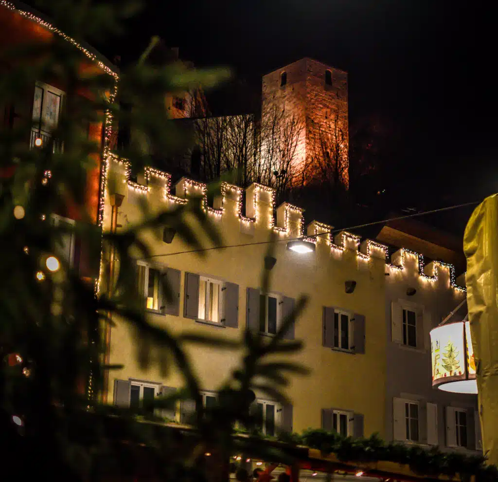 Bruneck's medieval castle tower lit up at night above the old town, framed by market lights and a painted lantern