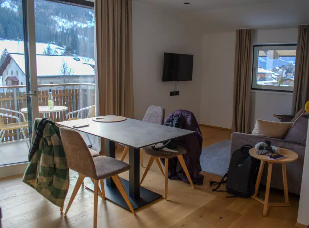 Open-plan living and dining area at Appartements Hofer with a dining table, sofa, wall-mounted TV, and balcony overlooking snowy Dobbiaco.