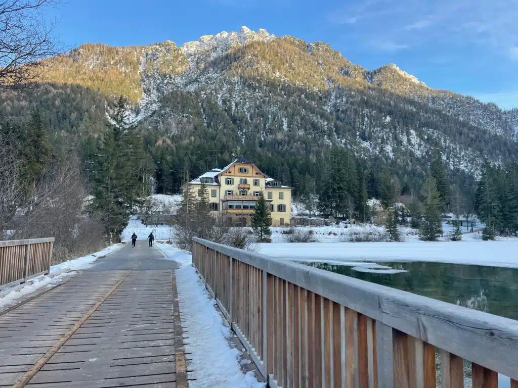 Snowy wooden bridge leading toward a historic hotel and Dolomite peaks at Lago di Dobbiaco, winter walk, Italy.