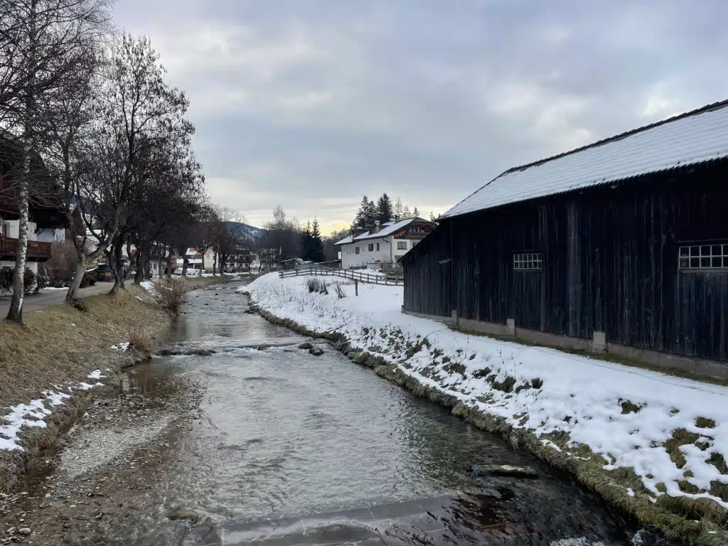 Snow-lined stream running through Villabassa (Niederdorf) past a dark timber barn and alpine houses in winter, Val Pusteria.